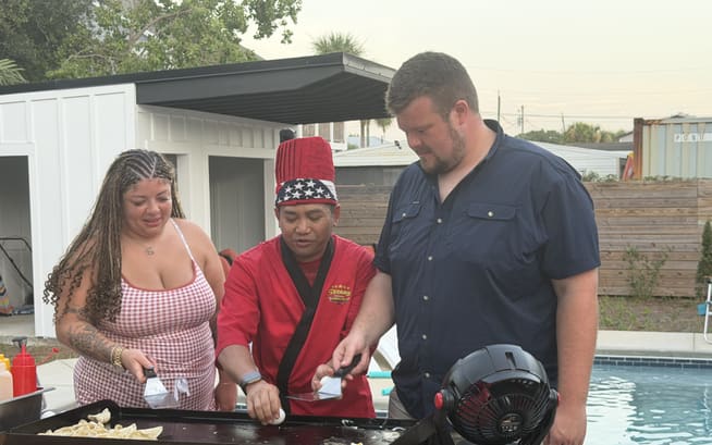 Chef in a red jacket cooking on the flat-top between two guests beside a pool at dusk