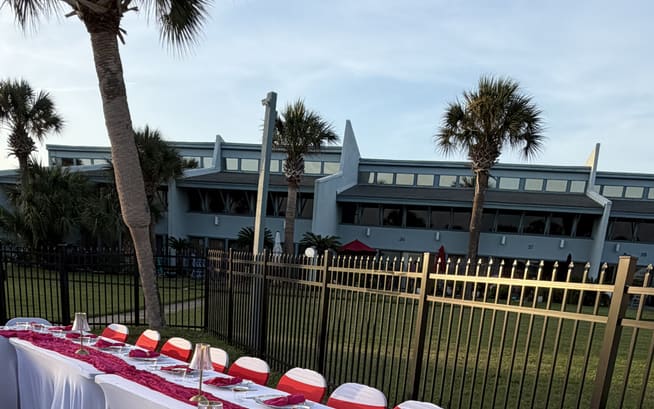 Long table dressed with a deep pink runner and gold place settings beside a pool, dark railing behind