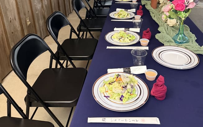 A long dinner table set with plated salads under a rental-house carport, folding chairs down both sides