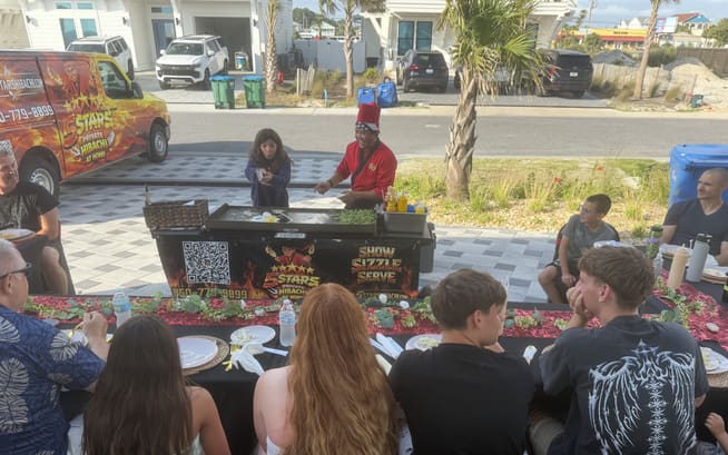 A group seated along a long table in a paved driveway with palms behind and the hibachi cart at the head
