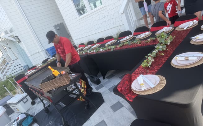 Chef working the flat-top beside a long table with a red runner outside a white clapboard rental house
