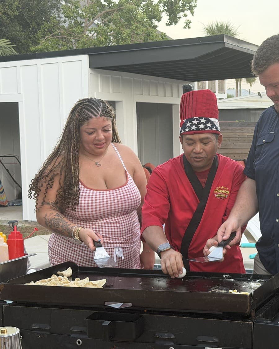 Chef in a red jacket cooking on the flat-top between two guests beside a pool at dusk