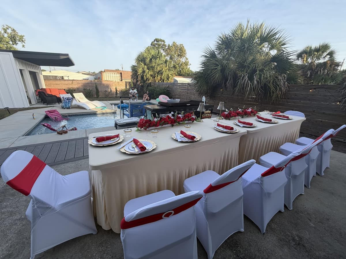 A long table dressed in white with red sashes and roses beside a pool, palms and low resort buildings behind it