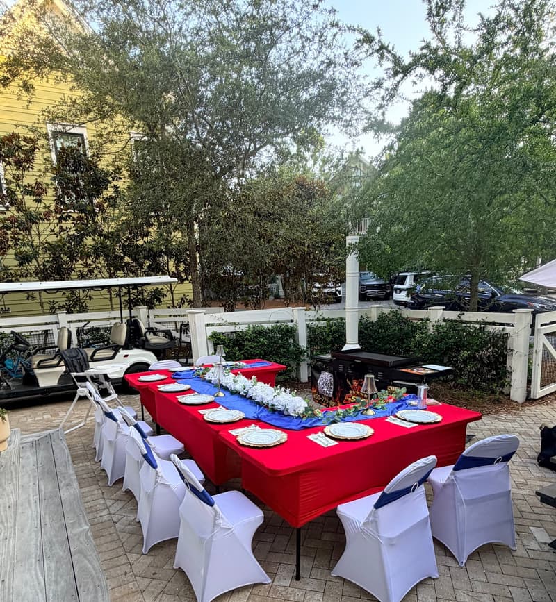 Long table laid on a brick paver courtyard beside a pastel cottage, white picket fence and a parked golf cart behind