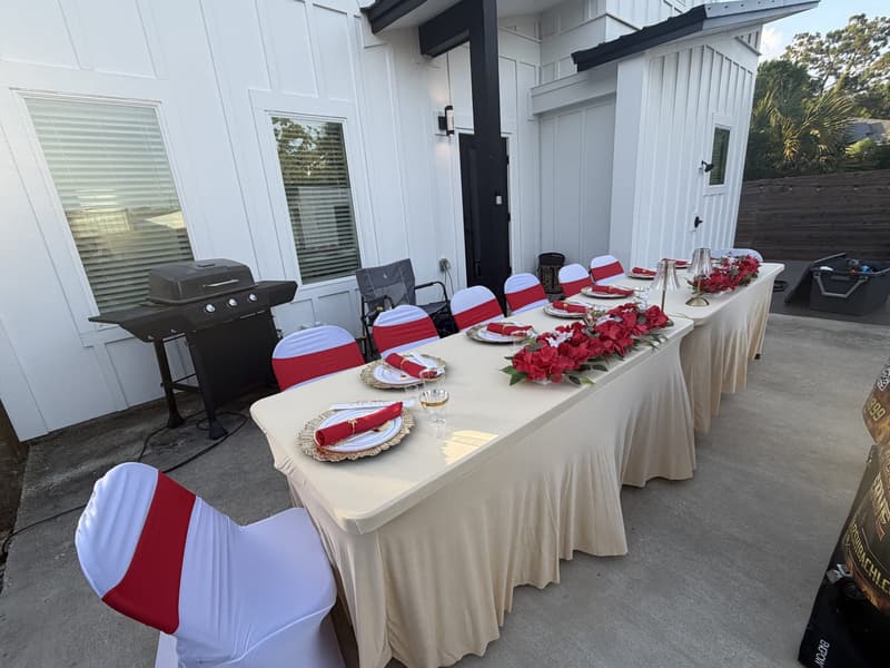 A long table with red roses and gold chargers set on a paved patio beside a modern white beach house, gas grill to one side