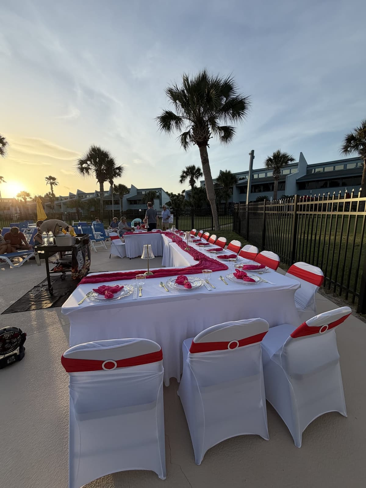 Long tables in white covers with pink runners set out along a pool deck at dusk, the grill at the far end