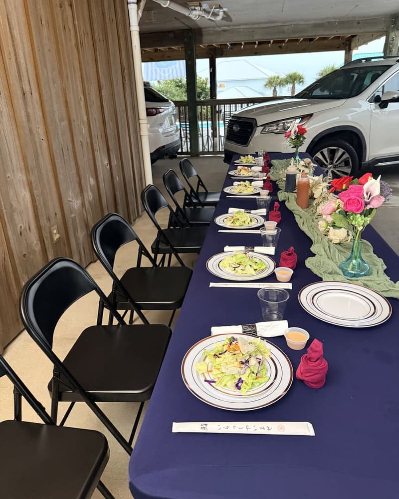 A long dinner table set with plated salads under a rental-house carport, folding chairs down both sides