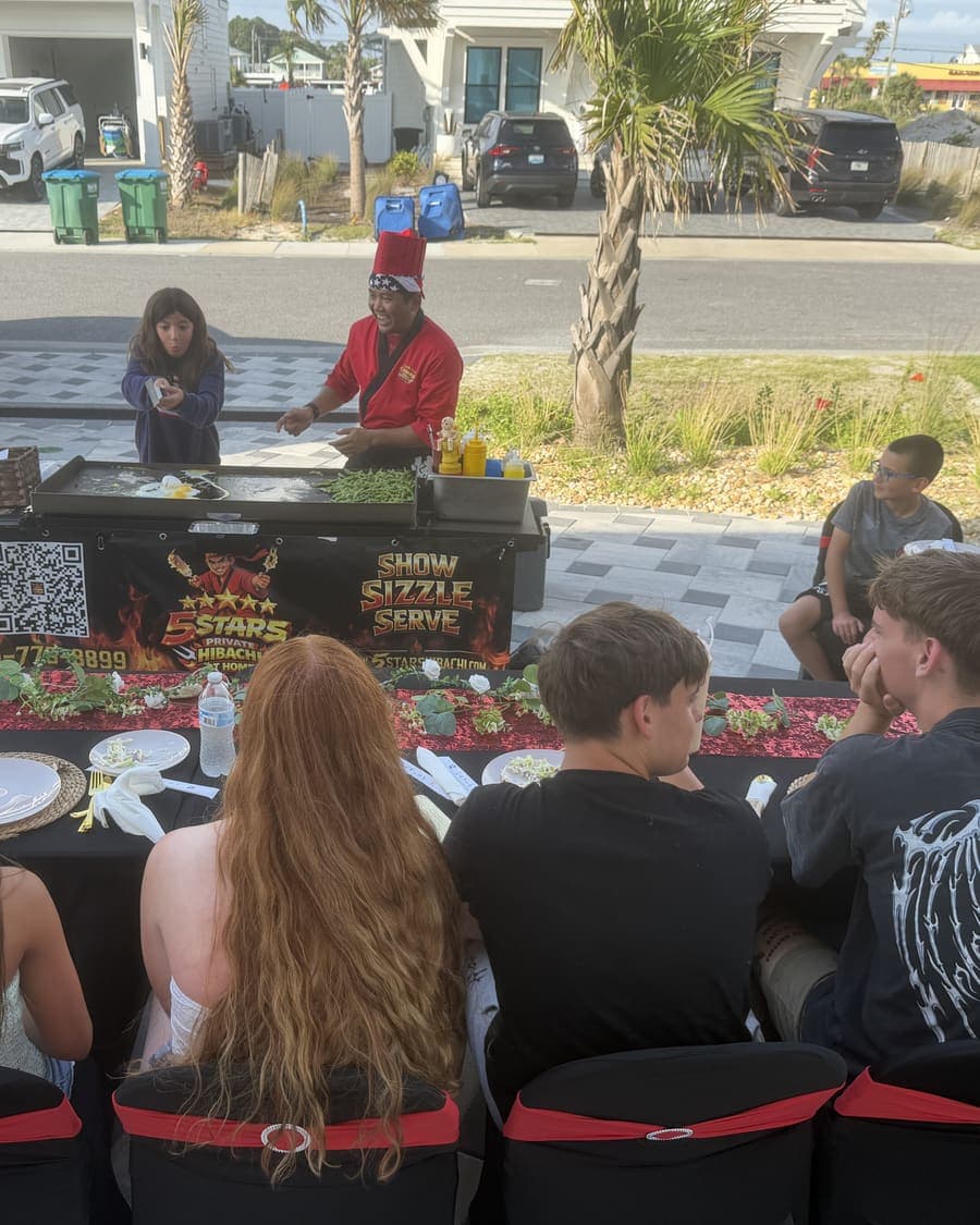 A group seated along a long table in a paved driveway with palms behind and the hibachi cart at the head