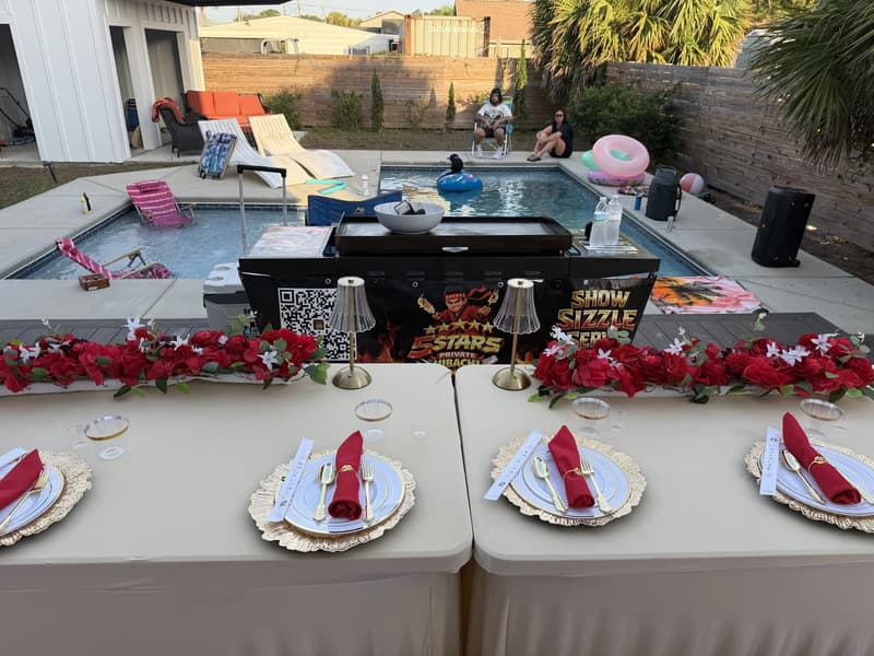 A long table with red roses and place settings beside a swimming pool, loungers along the far edge
