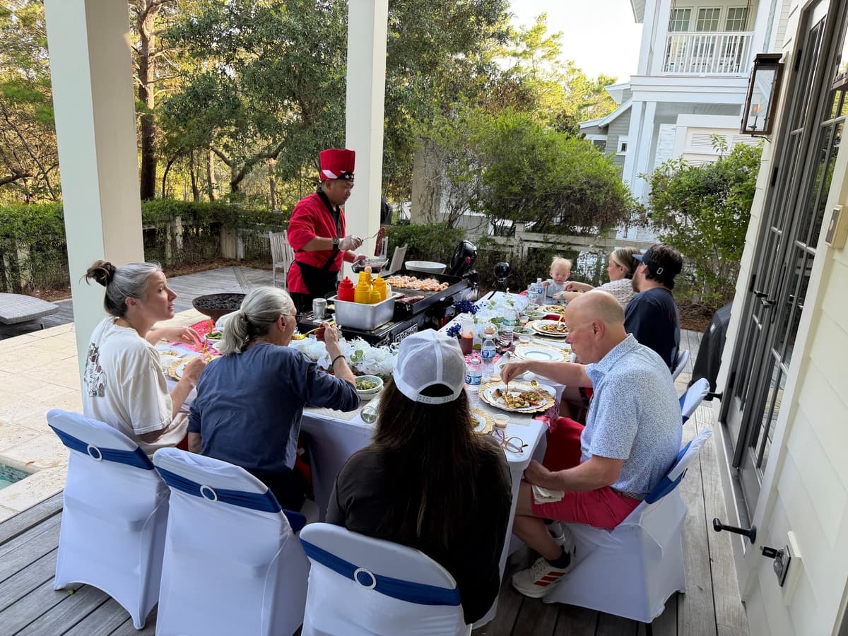 Guests seated the length of a long table on a shaded porch while the chef works the flat-top at the head