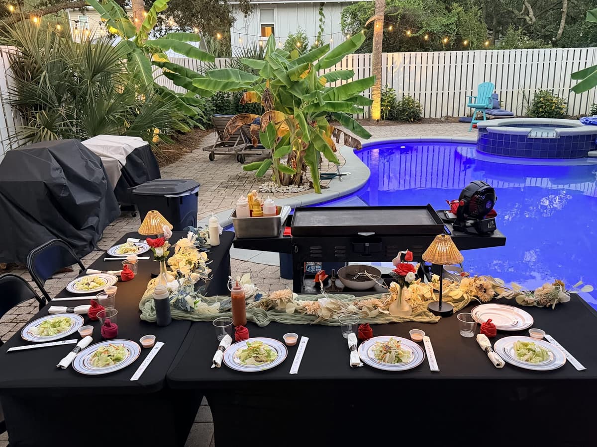 A long table set beside a lit swimming pool at dusk, the flat-top grill standing ready at one end