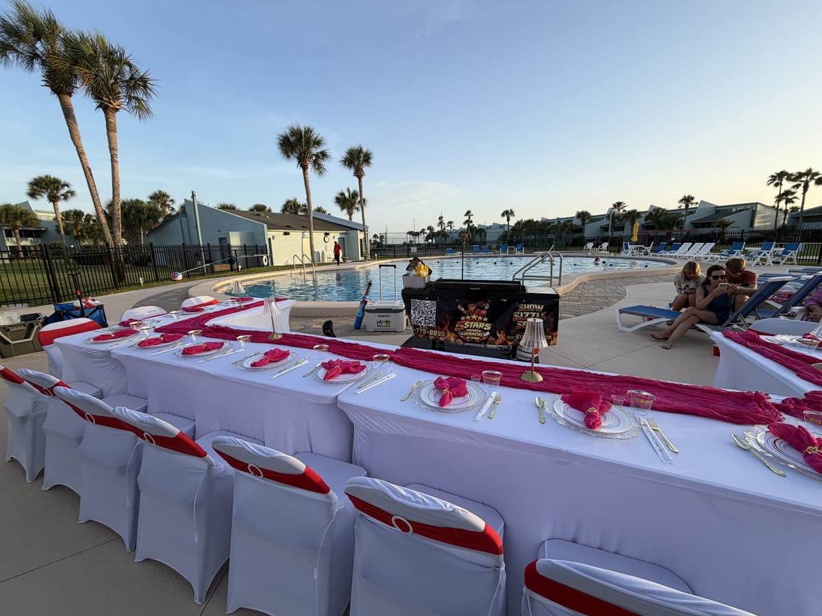 Long tables in white covers with pink runners set along a resort pool deck at dusk, the hibachi cart at the far end