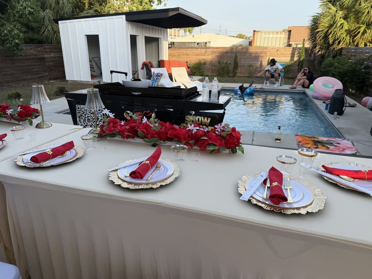 A table dressed with red roses and gold chargers on a paved terrace beside a pool, a modern white house behind it