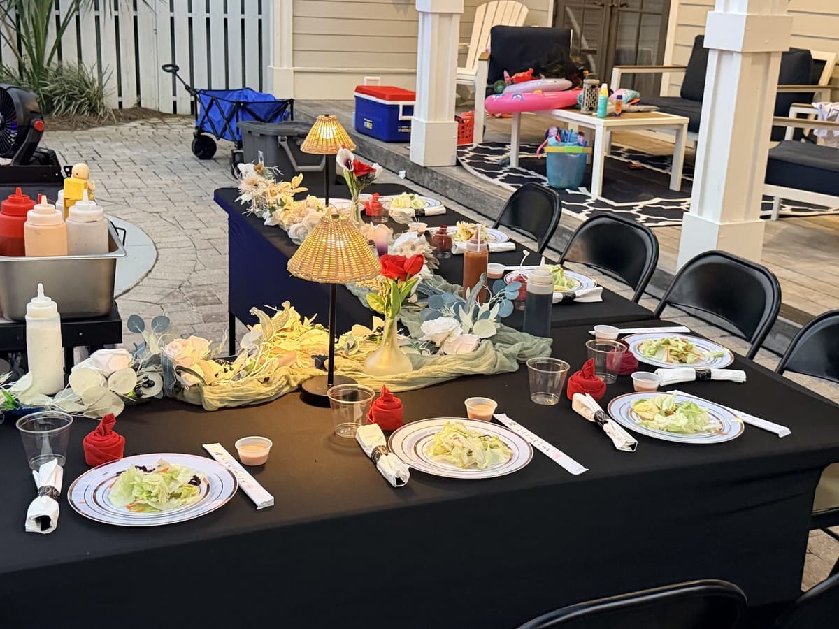 A long table lit with small shaded lamps and set with plated salads on a deck beside a cottage at dusk