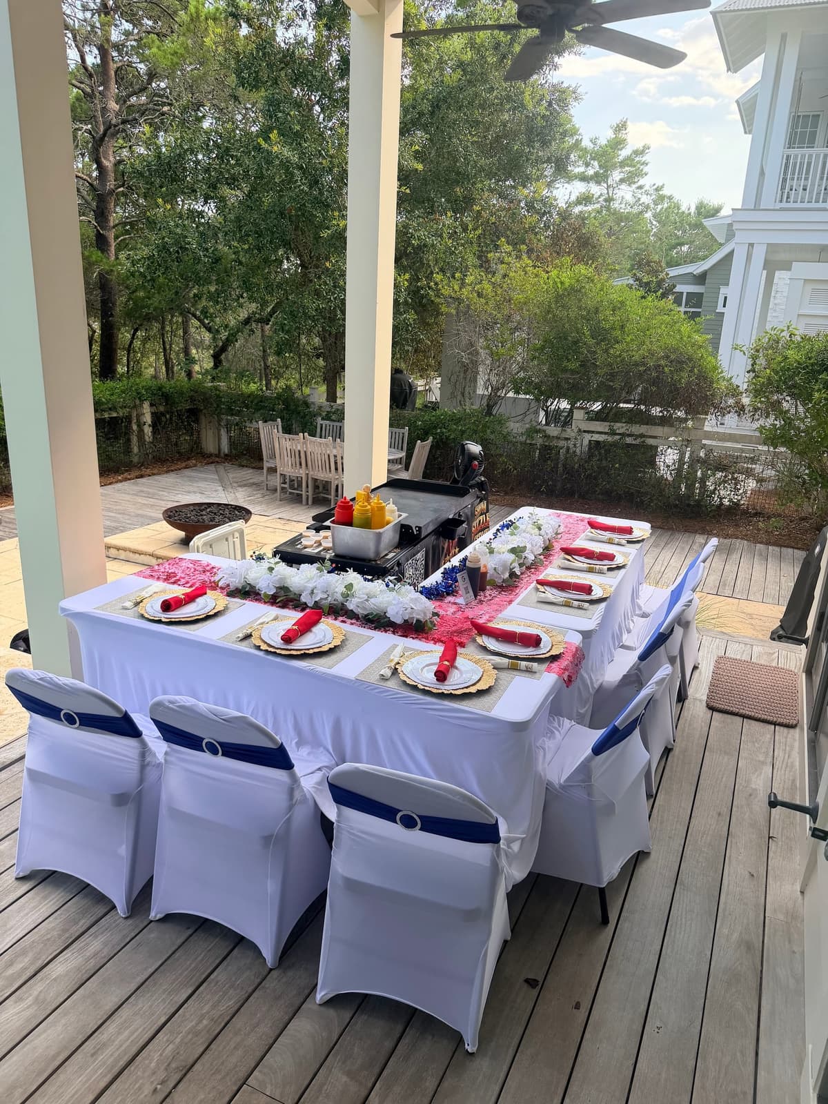Porch table laid with chargers, florals and a red napkin at every place, the grill station waiting at the head