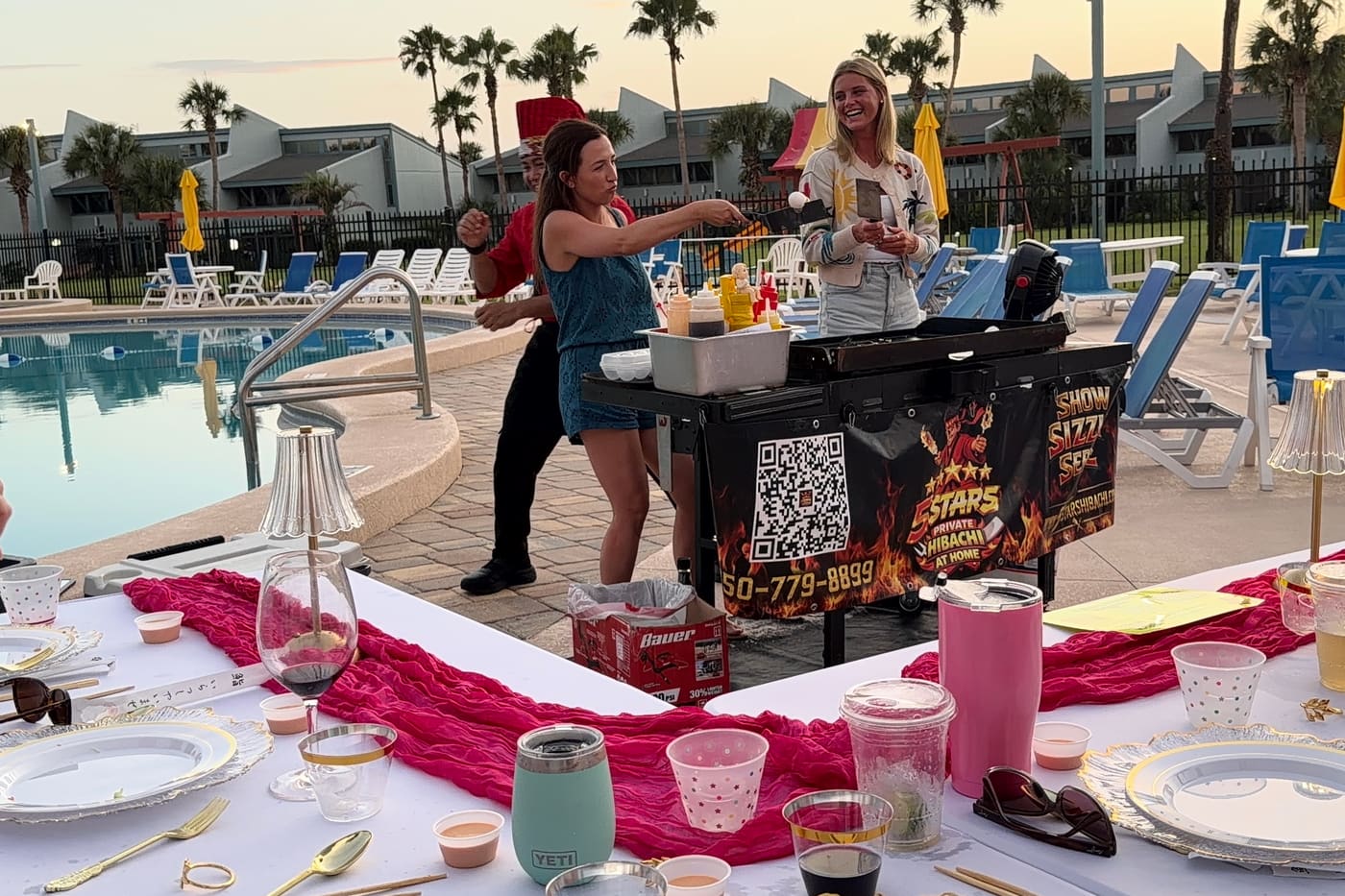 Two guests hugging beside the hibachi cart at a poolside table dressed with a pink runner