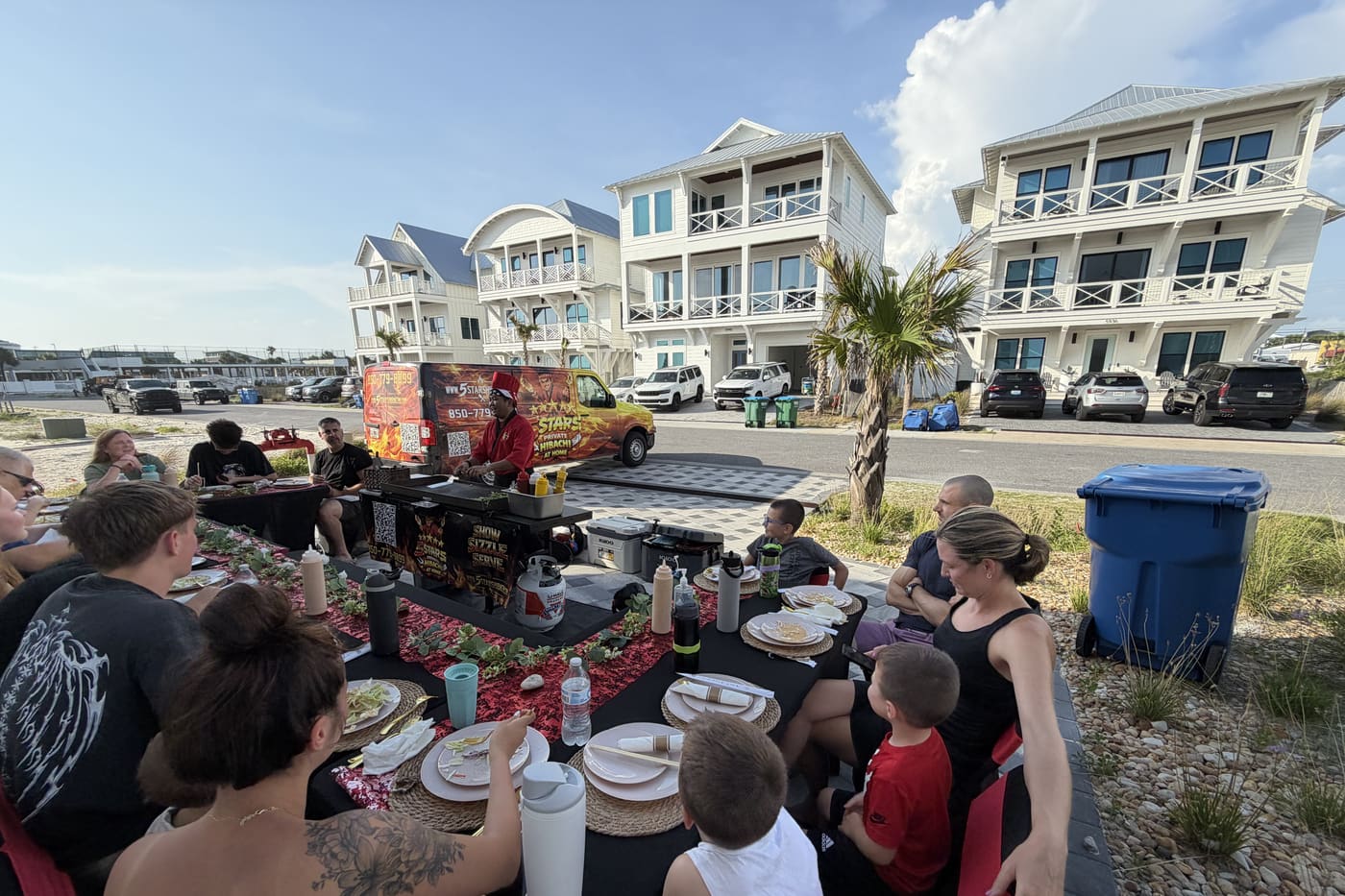 A family seated at a long table on a street-side setup between beach houses, chef cooking at the head