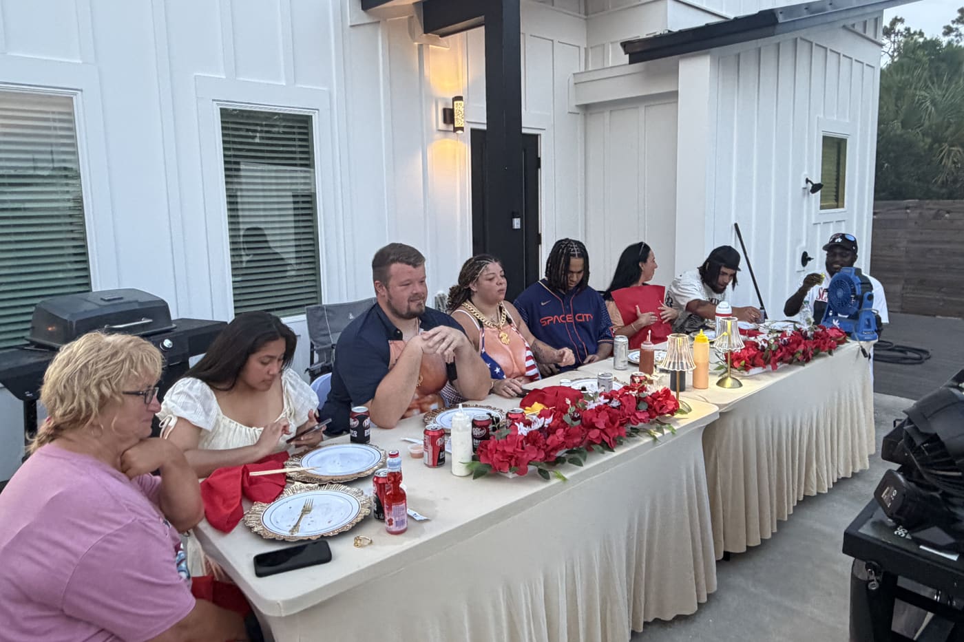 A large group seated down both sides of one long table beside a pool at a white rental house