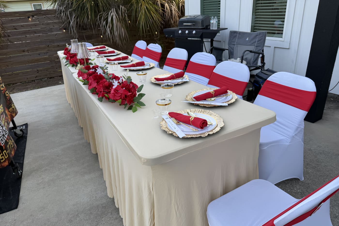 A long table run with red roses and white chair covers, set for a formal dinner outside a house